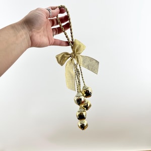 May include: Gold-colored decorative hanging bells with a gold bow and twisted rope. The bells are heart-shaped and hang in a vertical line. The bow is made of a shiny gold material. The rope is a twisted gold color.