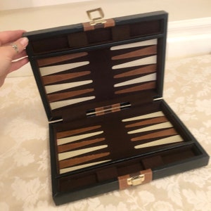 May include: A black leather backgammon game set with a brown felt playing surface and ivory and brown playing pieces. The set is closed and the lid is open.