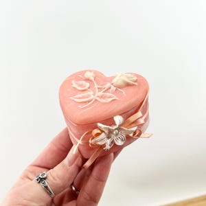 May include: A pink heart-shaped trinket box with a white rose and leaves design. The box is tied with a peach-coloured ribbon and a white flower.