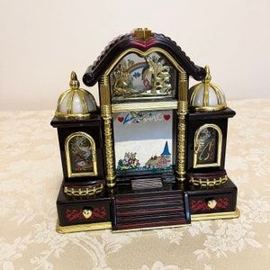 May include: A dark brown wooden musical jewelry box with gold accents. The box features a whimsical scene with a rainbow, a castle, and a bird. The box has a glass front and a small drawer on the bottom.