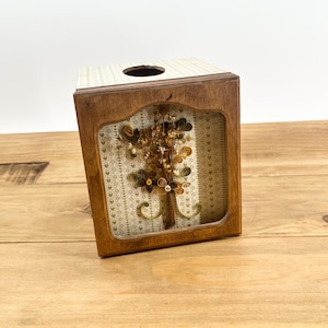 May include: A wooden tissue box cover with a clear window displaying a brown, yellow, and green paper flower arrangement. The tissue box cover has a white and green patterned background with small hearts.