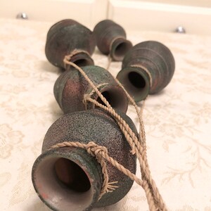 May include: A set of five small, brown, textured ceramic pots hanging from a thick, brown rope. The pots are tied together with the rope and are arranged in a vertical line.