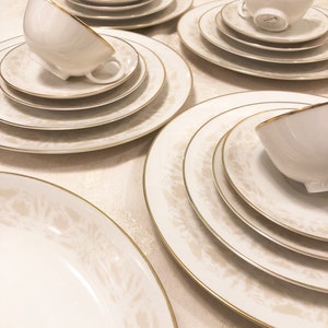 May include: Elegant white porcelain dinnerware set with gold trim. The set includes cups, saucers, and plates of various sizes, featuring a subtle floral pattern. The dishes are arranged on a white tablecloth.