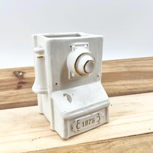 May include: A white ceramic telephone-shaped vase with gold accents. The vase has a rectangular body with a rotary dial and a handset. The base of the vase has the number "1876" in gold lettering. The vase is on a wooden surface.