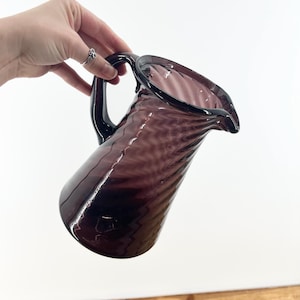 May include: A vintage purple glass pitcher with a handle and a spout. The pitcher has a textured, swirled design and is held against a white background. The pitcher is a deep shade of purple.