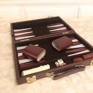 May include: A brown leather carrying case for a backgammon set. The case is open, revealing the game board, dice, and playing pieces. The board is made of wood and has a burgundy and white checkerboard pattern. The dice are white with black dots. The playing pieces are white and burgundy.
