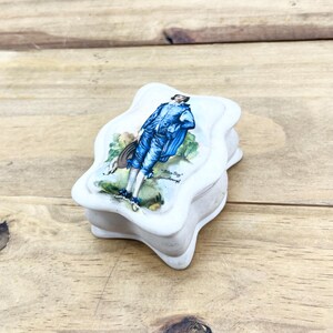 May include: White ceramic trinket box with a lid depicting a painting of a man in a blue suit, titled "Blue Boy" by Gainsborough. The box has a distinctive, wavy shape and is on a wooden surface.