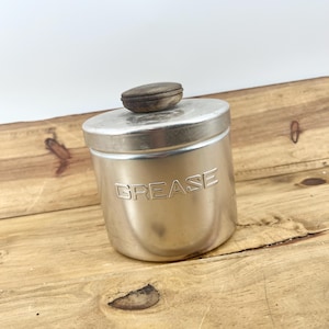 Peut inclure: Un récipient à graisse cylindrique en métal argenté doté d'un couvercle assorti avec un bouton en bois brun foncé. Le mot GREASE est gravé en lettres capitales sur la face avant du récipient, qui présente une finition métallique lisse.