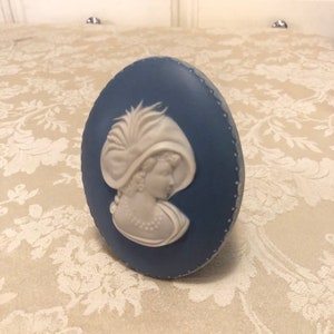 May include: Oval blue ceramic plaque with a white bas-relief profile of a woman in a feathered hat and pearl necklace. The plaque has a dotted border. The background is patterned.