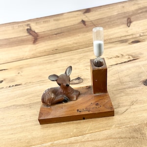 May include: A wooden base featuring a small, brown, resting deer figurine and a vertical wooden block holding a glass hourglass filled with white sand. The base has text printed on it that reads "FOR THE SCHULTZ'S COLORADO".