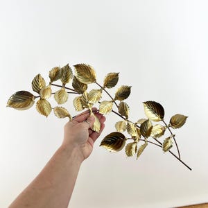 May include: A decorative gold-colored metal leaf branch with detailed leaves. The leaves have a textured surface and are arranged along thin, brown stems. The branch is held by a hand against a white background, showcasing the metallic finish.