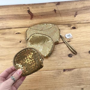 May include: Three gold mesh coin purses of varying sizes are displayed on a wooden surface. The smallest purse is held by a hand with a silver ring. The purses have zipper closures and a wrist strap.