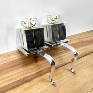 May include: A pair of silver stocking holders shaped like gift boxes with gold bows. The holders are on a wooden surface.