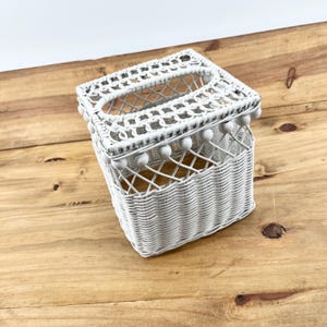 May include: White wicker tissue box cover with a rectangular opening on top. The cover has a decorative border with small white ball accents. The basket weave design is visible on the sides and bottom of the box.