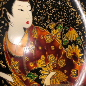 May include: A black ceramic plate with a gold and red painted design of a woman in a traditional Japanese kimono. The woman is holding a fan and has a gold and red floral design on her kimono.