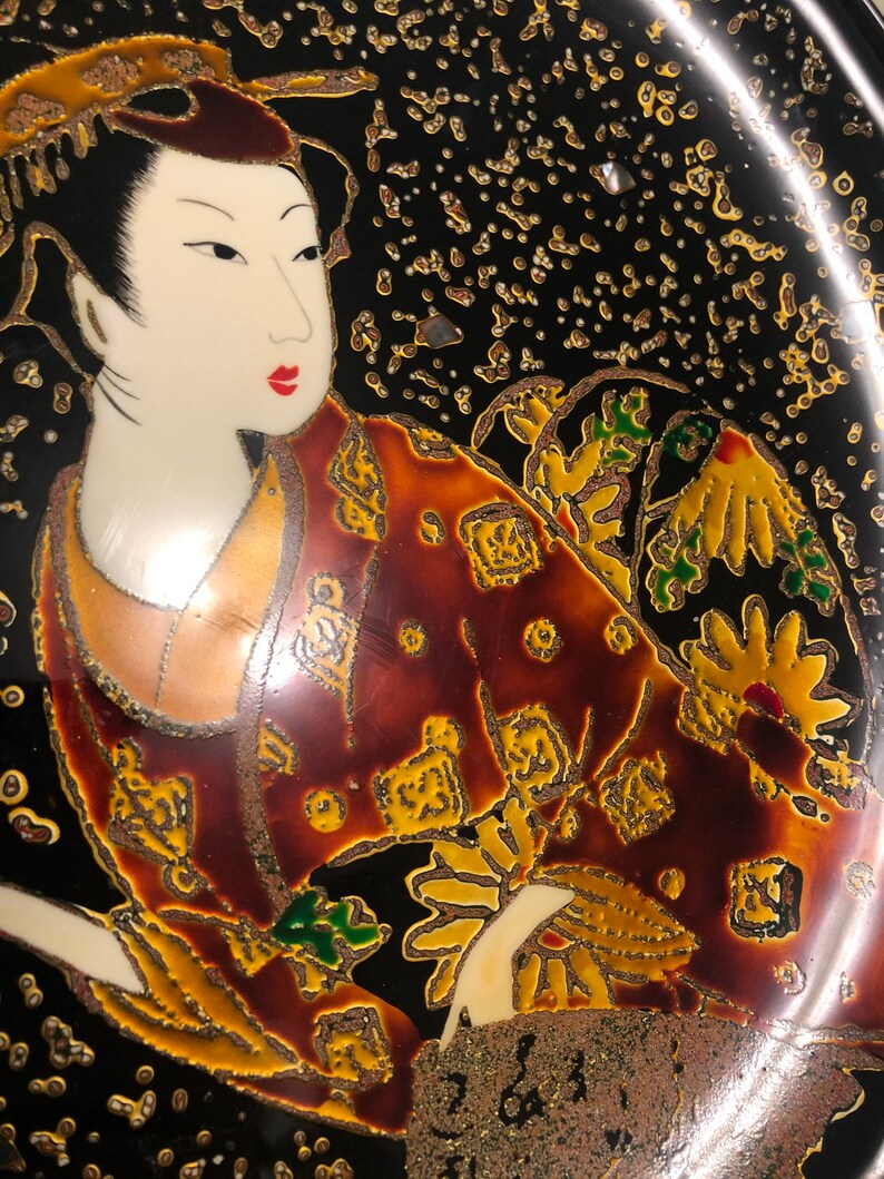May include: A black ceramic plate with a gold and red painted design of a woman in a traditional Japanese kimono. The woman is holding a fan and has a gold and red floral design on her kimono.