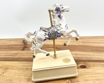 May include: A white carousel horse figurine with floral accents in purple and blue. The horse is mounted on a cream-colored music box with a gold-colored pole. The figurine is set on a wooden surface.