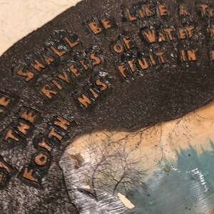 May include: A brown, textured, rectangular sign with the text "I SHALL BE LIKE THE RIVERS OF WATER FOYTH MISS FRUIT IN T" printed in gold lettering. The sign has a faded image of a forest scene in the background.