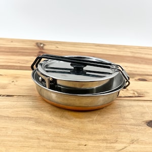 May include: A stainless steel camping cook set with a lid and folding handle. The set includes a pot and a pan, perfect for outdoor cooking. The set is compact and designed for easy storage and transport.