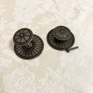 May include: Two antique bronze cabinet knobs with a circular design. Each knob features a smaller, raised center circle surrounded by a larger, flat, decorative disc. One knob has a screw attached. The knobs are on a cream-colored floral patterned background.