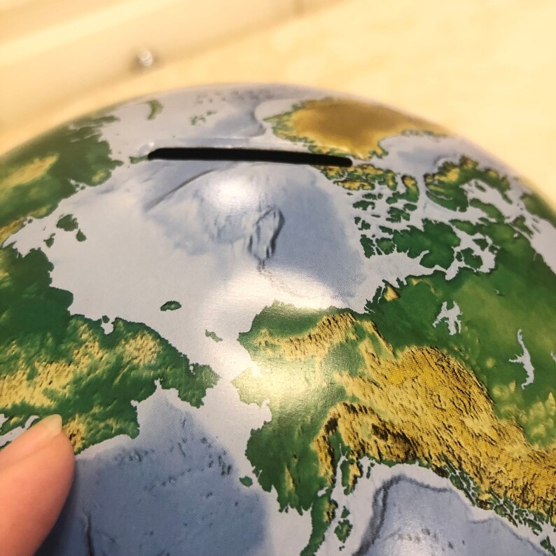 May include: A blue and green globe piggy bank with a slot on the top for coins. The globe has a detailed map of the world with continents and oceans.