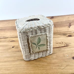 May include: A square white wicker tissue box cover featuring a braided trim along the top edge and a centered square ceramic tile inlay. The tile is off-white with a painted illustration of three green leaves in the center of the surface.