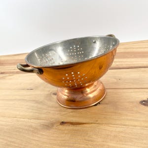 May include: A copper colander with a silver interior and small drainage holes. The colander has two small handles and sits on a copper base. The colander is on a wooden surface.