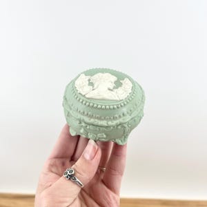 May include: A small, round, pale green trinket box with a white cameo design on the lid. The box has an ornate design with a beaded edge and decorative feet. The cameo features a woman's profile. A ring is visible on a hand holding the box.