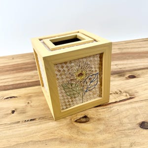 May include: A square, light yellow wooden tissue box with a woven, beige panel featuring a sunflower embroidery. The top has a rectangular opening. The box sits on a wooden surface.