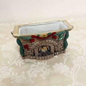 May include: A ceramic planter shaped like a brick fireplace with a green garland and the words "Merry Christmas" on the mantle. The fireplace has a brown brick exterior, a black firebox, and a red and white stocking hanging from the mantle.