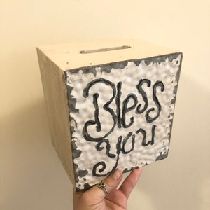 May include: White wooden box with a metal panel on the front. The metal panel has a textured surface and the words "Bless you" written in black.