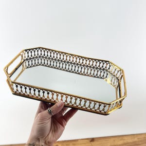 May include: A gold-colored, octagonal decorative tray with a mirrored surface. The tray has a delicate, openwork design around the edges and handles on each end. The tray is held up against a white background.
