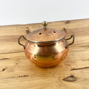May include: A copper potpourri container with a rounded body, two side handles, and a domed lid featuring small ventilation holes. A brass plaque is attached to the front of the vessel with the word POTPOURRI engraved in capital letters.