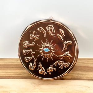 May include: A copper-colored, round wall hanging featuring zodiac symbols around a central sunburst design. The word "MIRRO" is visible in the center. The hanging has a decorative, ridged edge and a small loop for hanging.