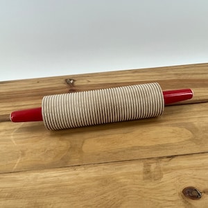 May include: A beige rolling pin with red handles. The rolling pin has a ridged surface.