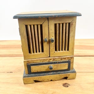 May include: A small, vintage wooden cabinet with a distressed yellow finish and dark blue trim. The cabinet features two doors with vertical slats and a small drawer below. The hardware includes small, round knobs.