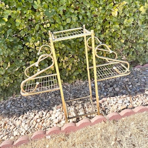 May include: A vintage gold metal plant stand with three wire shelves and decorative curved hanger-like arms. The stand is designed to hold plants and other decorative items. The stand is outdoors, set against a backdrop of green foliage and a bed of small rocks.