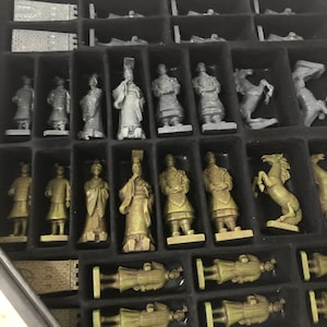 May include: A set of chess pieces in a black box. The pieces are made of painted metal and are in the style of the Terracotta Army. The pieces are arranged in rows on a black felt surface.