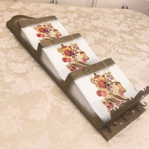 May include: A vintage metal wall-mounted mail holder with three compartments. Each compartment features a white background with a colorful illustration of fruit and teapots. The holder is painted in a distressed gold finish.