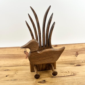May include: A wooden duck-shaped desk organizer with tall, curved reeds in the back. The duck has a detailed beak, wings, and legs with small round feet. The organizer is made of a light brown wood and sits on a wooden surface.