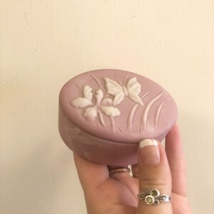 May include: A small, oval-shaped trinket box made of pale pink ceramic. The box is decorated with a raised design of white butterflies and flowers.