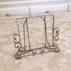 May include: A silver metal cookbook stand with decorative scrollwork details. The stand is designed to hold a cookbook open for easy reading while cooking.