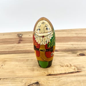 May include: A hand-painted wooden Santa Claus figurine. The figure has a light brown face, a white beard, and is wearing a red coat with a gold belt. A green Christmas tree design is on the side. The figurine is approximately 4 inches tall.