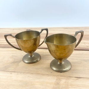 May include: A set of two gold-toned metal creamer and sugar bowls with handles. The bowls are on a wooden surface.