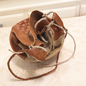 May include: A brown leather drawstring bag with a woven design and a beige fabric lining. The bag has a long brown leather strap.