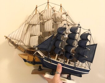May include: Two wooden model ships with white and blue sails. The larger ship has blue sails and the smaller ship has white sails with blue stripes. The ships are on a wooden base.