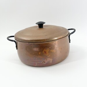 May include: A copper pot with a black lid and handles. The pot has a rounded shape and a slightly tarnished finish.