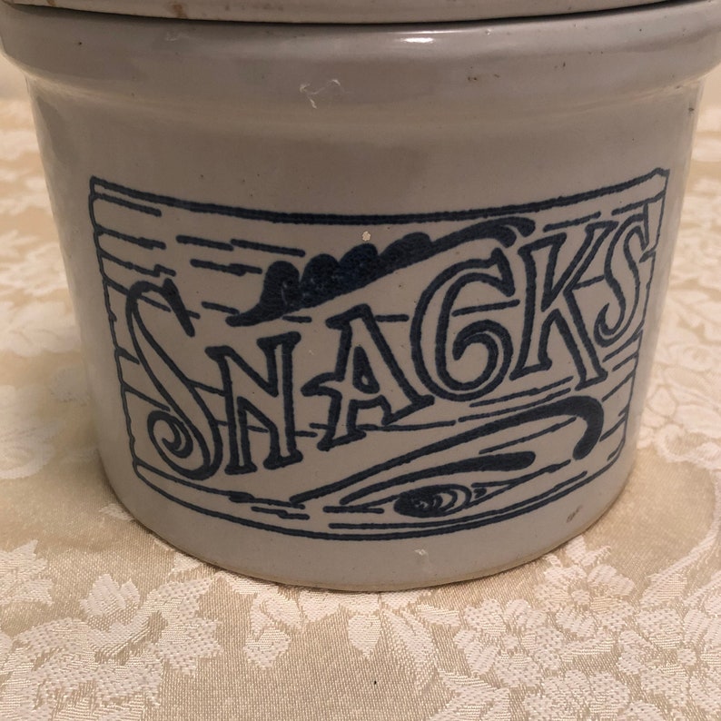 May include: A white ceramic crock with a blue and white design. The crock has the word "SNACKS" written on it in a decorative font.