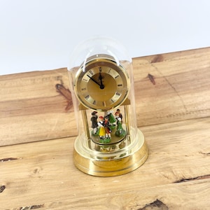 May include: A gold-toned clock with a clear dome, featuring a clock face with black hands and Roman numerals. Inside, miniature figures dance on a rotating platform. The clock sits on a gold base, creating a vintage aesthetic.