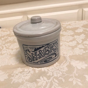 May include: A white ceramic jar with a blue and white design that says "SNACKS". The jar has a lid and is perfect for storing snacks.